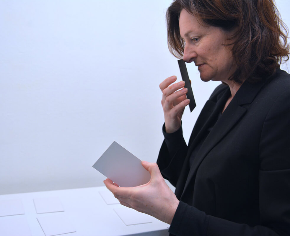 画像：白い空間の中で小さな展示物を手に取り、匂いを嗅いでいる女性の様子