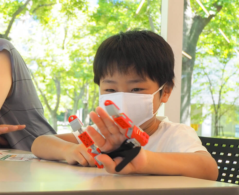 画像：LEGOブロックを組み立てて第６の指の機械を作り、自分の手に取りつけている男の子の様子