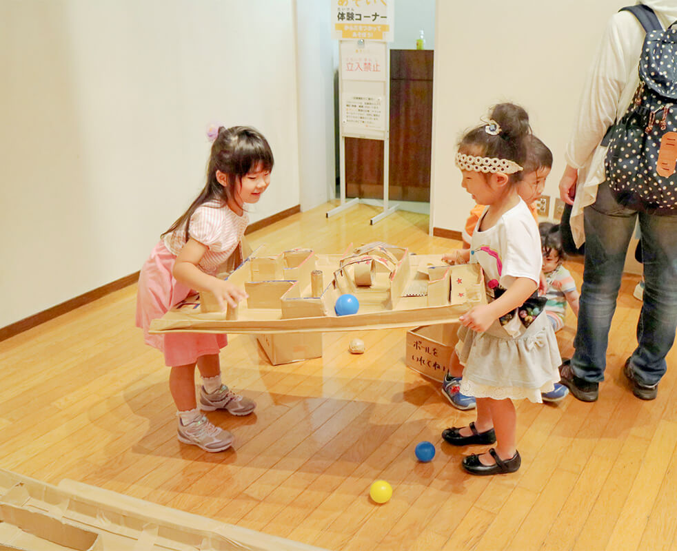 画像：段ボールで作られた小さな迷路を両側から抱え、その上のボールを転がす2人の女の子の様子