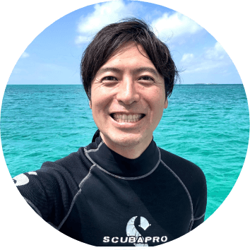 ウェットスーツ姿の男性が青い海をバックに笑顔で自撮りしている写真。