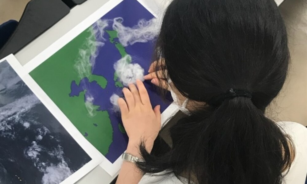 台風の雲を再現するワークショップの様子。女性が日本地図の上に綿を使った雲の模型を配置している。
