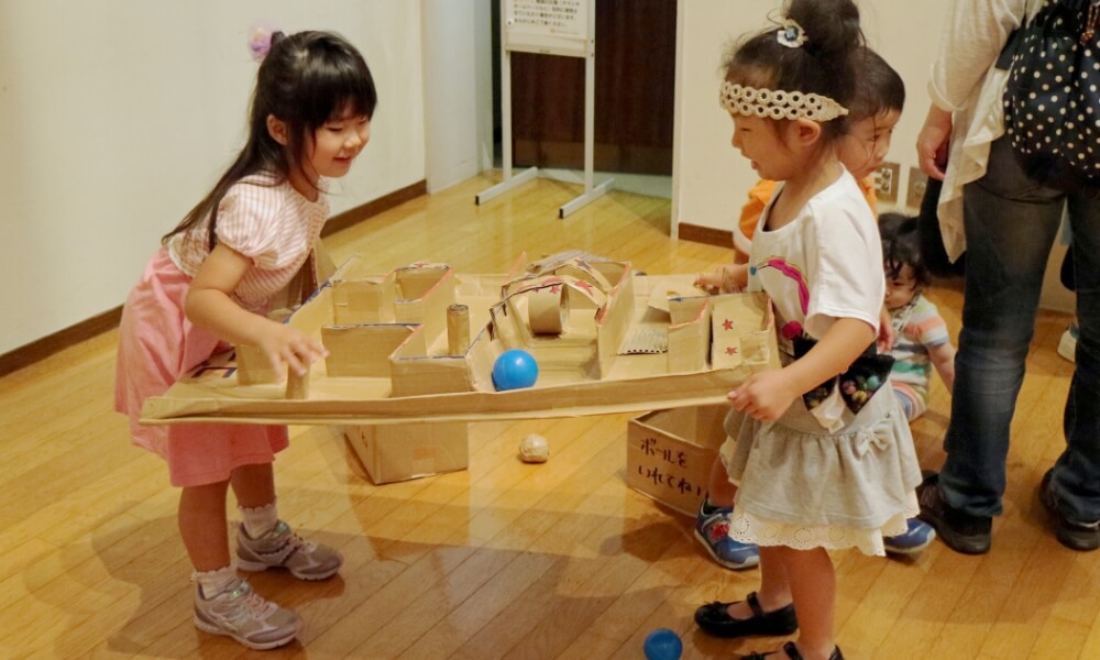 手作りの段ボール迷路でボールを転がしながら夢中で遊ぶ子どもたち
